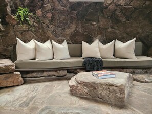 A stone wall, a beige sofa with white cushions, a patterned throw, and a book on a stone table.