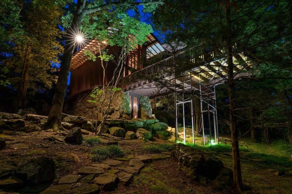 A wooden cabin with a metal staircase and a glass roof, surrounded by rocks and trees.