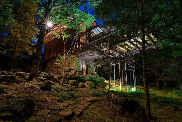 A wooden cabin with a metal staircase and a glass roof, surrounded by rocks and trees.