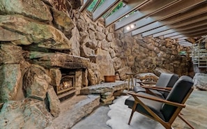 Una chimenea de piedra con fuego, rodeada por un muro de piedra y una zona de estar con sillones.