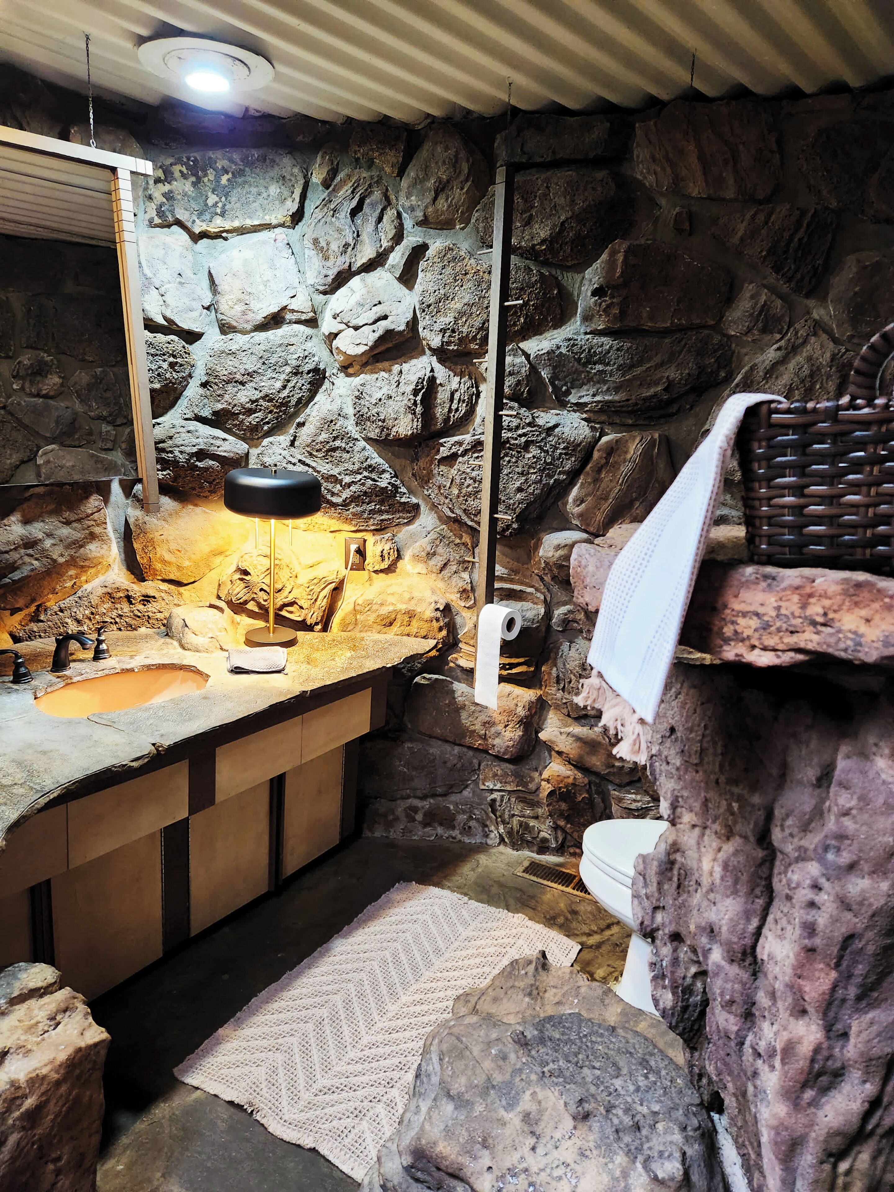 Un baño con pared de piedra, un lavamanos, un espejo, una toalla y una cesta.