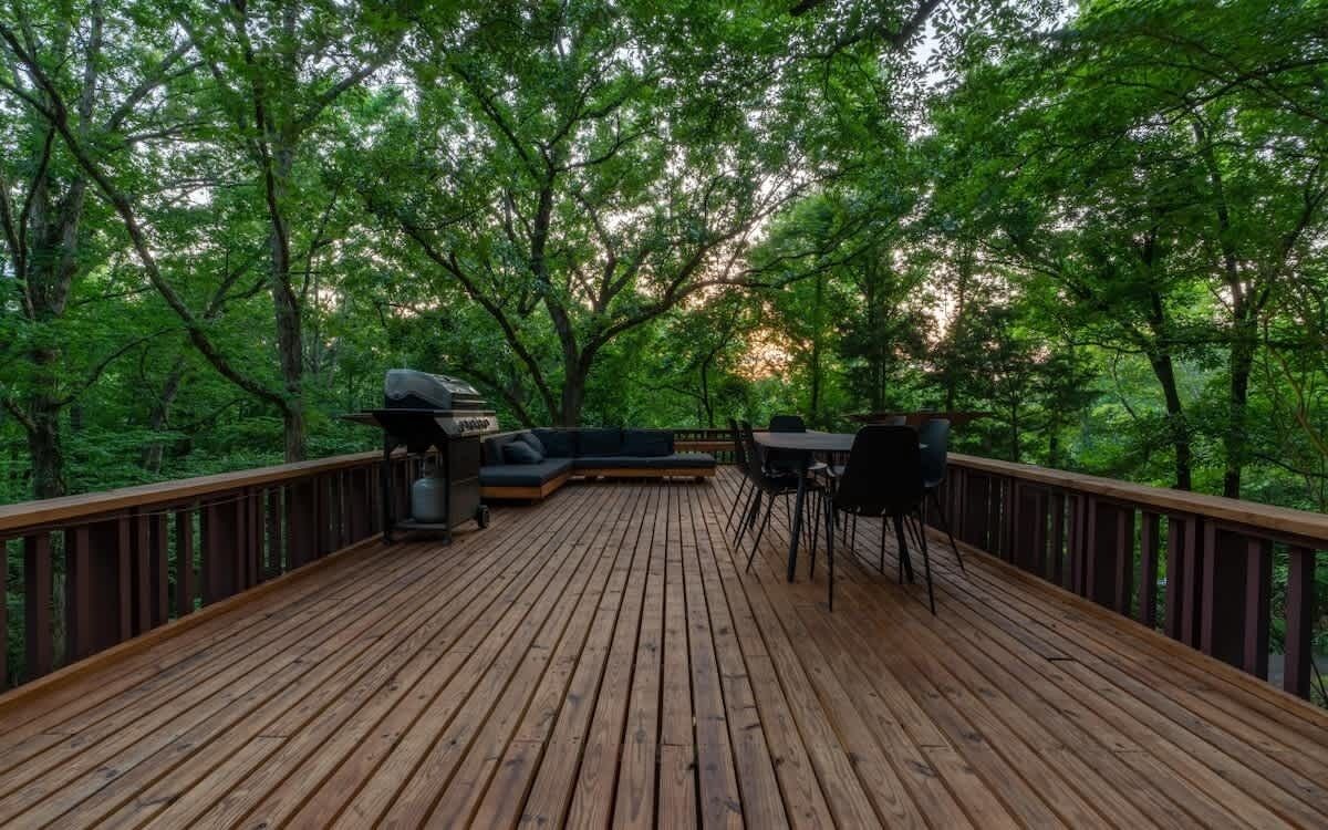 Una terraza de madera con una parrilla y zona de estar, rodeada de árboles.