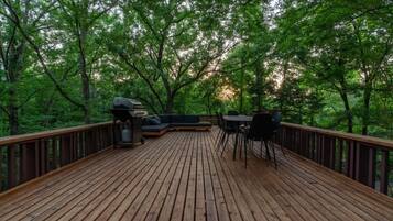 Una terraza de madera con una parrilla y zona de estar, rodeada de árboles.