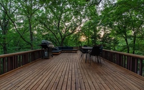 Une terrasse en bois avec un barbecue et un espace salon, le tout entouré d’arbres.