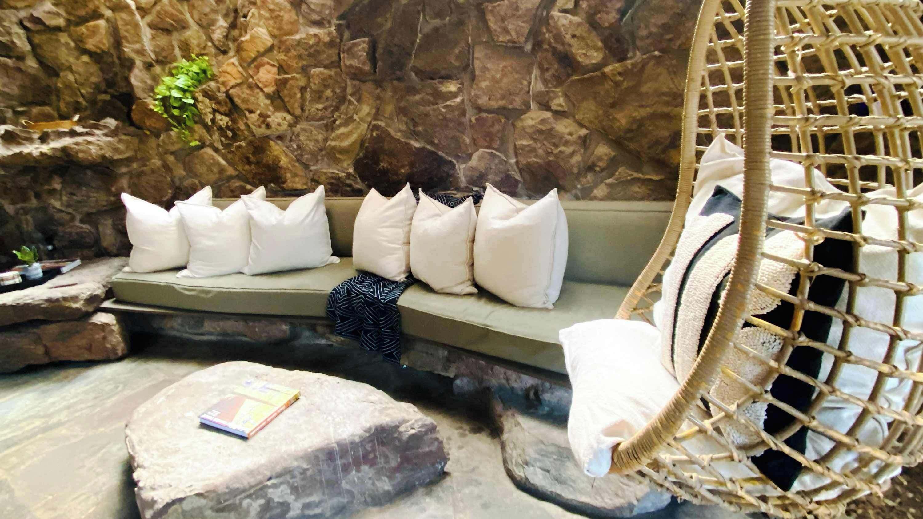 A stone wall, a cushioned bench with white pillows, a hanging chair, and a small plant.