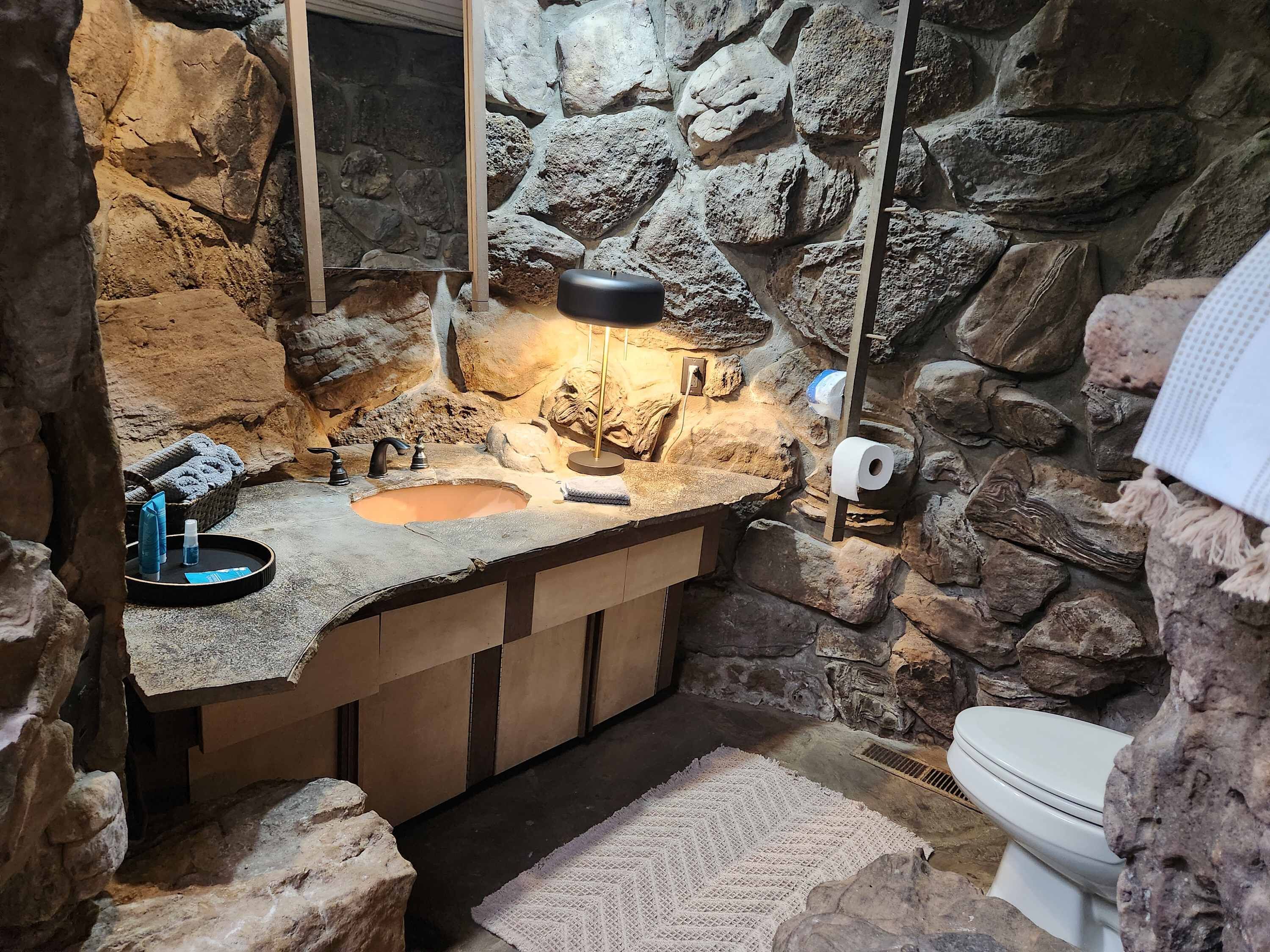 A bathroom with a stone wall, a sink with a mirror, a toilet, and a small rug.