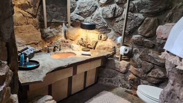 A bathroom with a stone wall, a sink with a mirror, a toilet, and a small rug.