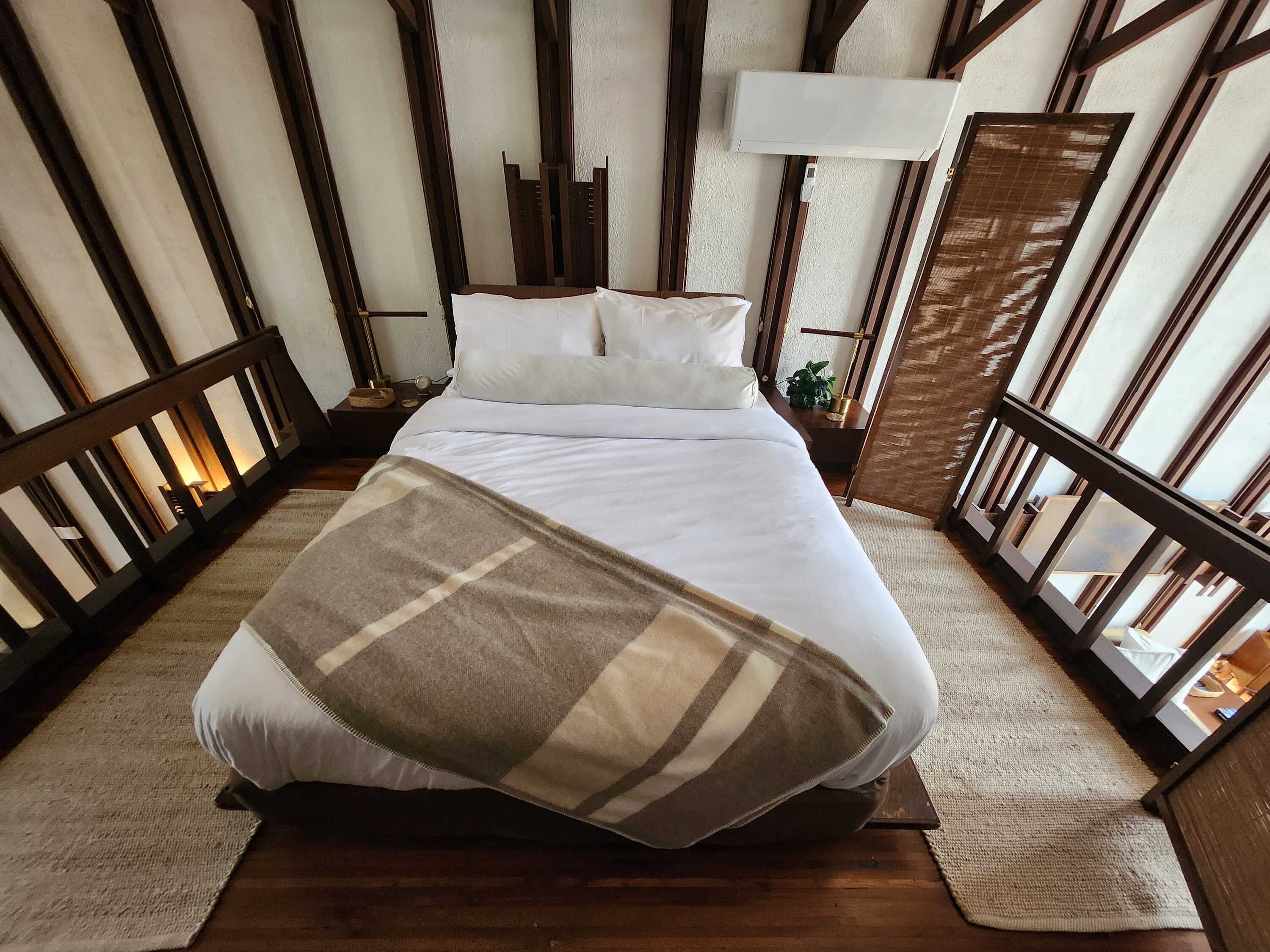 Un lit avec une literie blanche et une couverture à carreaux, un plancher en bois et une table de chevet avec une lampe.