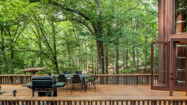 Une terrasse en bois avec un barbecue et des chaises, entourée d’arbres.