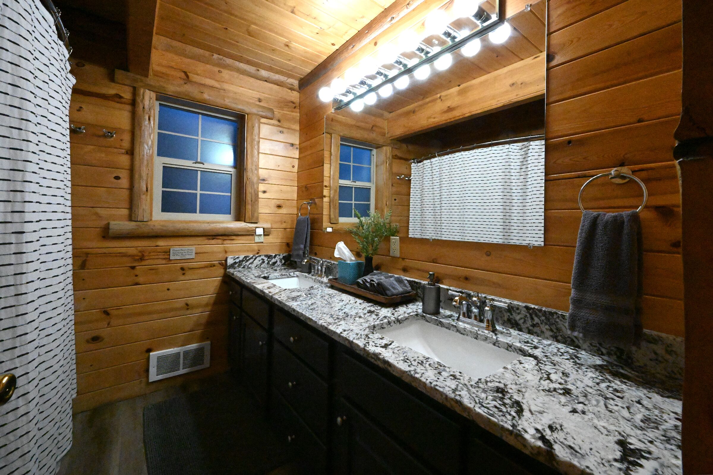 A wooden cabin bathroom with a granite countertop, two sinks, a mirror, and a towel ring.