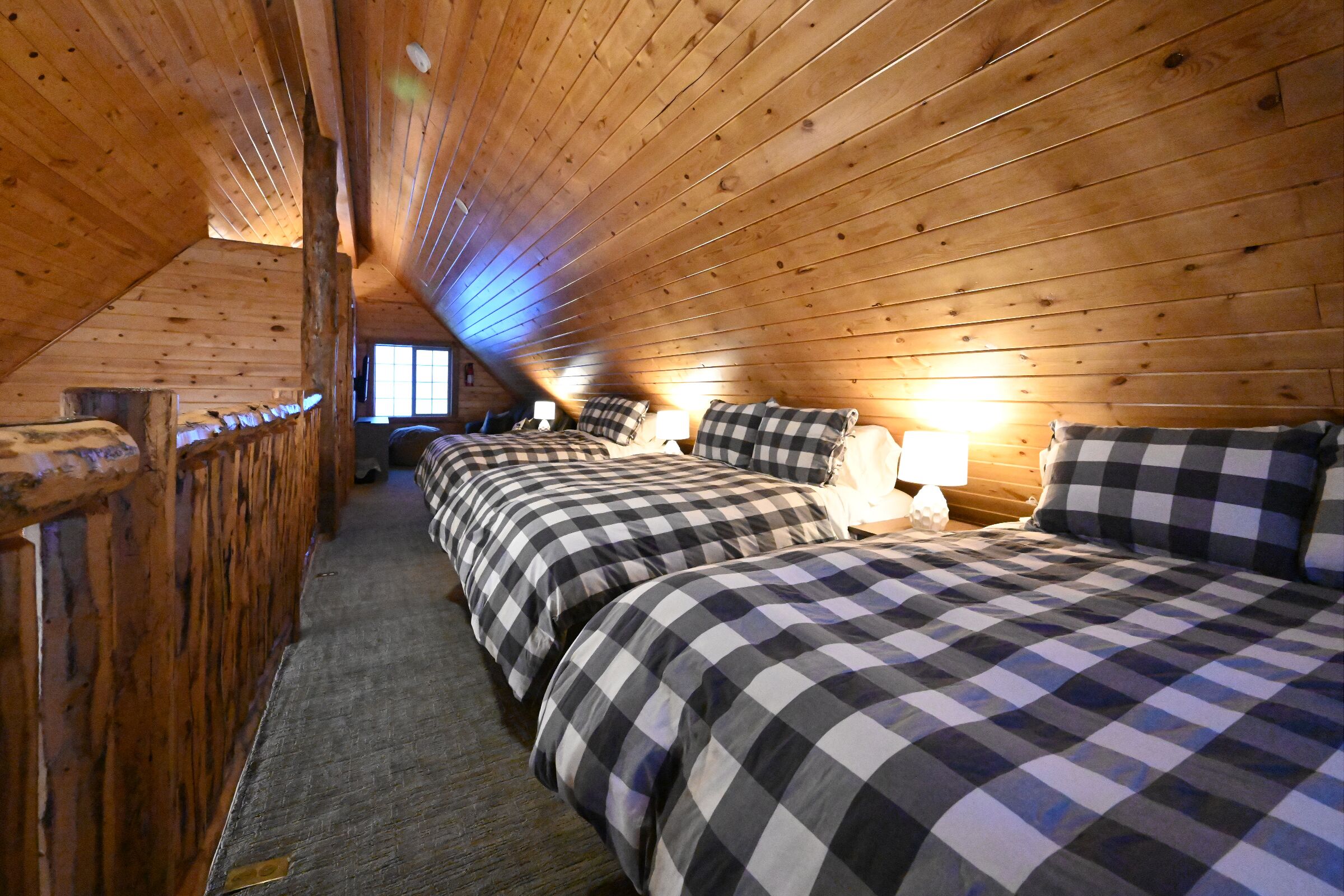 A cozy attic bedroom with wooden walls and sloped ceilings, featuring three beds with checkered bedding and bedside lamps.