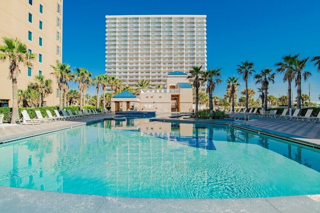 Piscina con sillas de descanso y palmeras, rodeada por un edificio alto de hotel.
