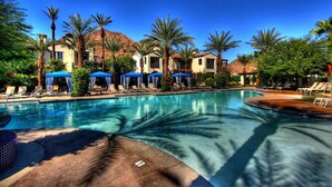 A swimming pool with lounge chairs and umbrellas, surrounded by palm trees and buildings.