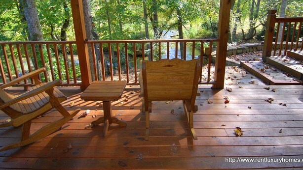 A wooden deck with a table and two chairs, surrounded by a wooden railing, overlooking a forested area.
