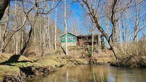A rustic cabin with a wooden deck, surrounded by a dense forest and a calm river.