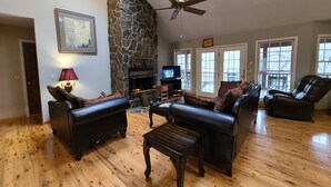 A spacious living room with a stone fireplace, leather sofas, a wooden coffee table, and a ceiling fan.