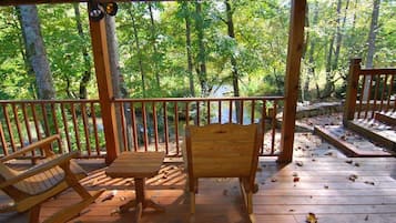 A wooden deck with a table and two chairs, surrounded by a wooden railing, overlooking a forested area.