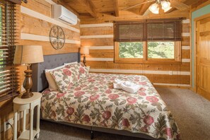 A wooden cabin bedroom with a bed, two bedside lamps, a window with blinds, and an air conditioning unit.