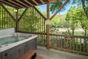 A wooden deck with a hot tub, wooden railings, and a view of a lake surrounded by trees.