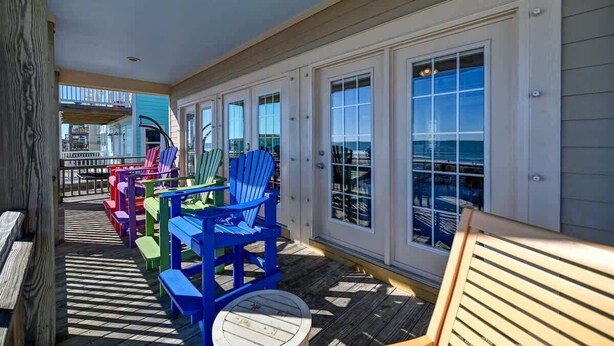A covered outdoor area with colorful Adirondack chairs and a wooden bench, a wooden deck, and a view of the ocean through large windows. - This' L Do - Oceanfront - Upcycled Vintage Beach Cottage (North Topsail Beach)