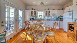 A modern kitchen with white cabinets, a dining table with chairs, and a view through large windows. - This' L Do - Oceanfront - Upcycled Vintage Beach Cottage (North Topsail Beach)