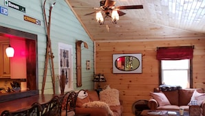 Ein gemütliches Blockhaus-Interieur mit Holzbalkendecke, einem Essbereich mit barähnlicher Einrichtung, einer Couch mit Decke und einem Couchtisch mit Gesellschaftsspielen.