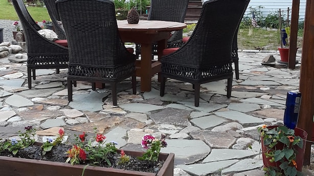 Eine Außenpatio mit Steinboden, einer Holz-Pergola, Korbstühlen, einem Holztisch und einem Blumenbeet.