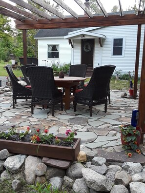 Eine Außenpatio mit Steinboden, einer Holz-Pergola, Korbstühlen, einem Holztisch und einem Blumenbeet.