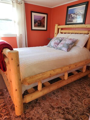Ein hölzernes Bett mit weißer Bettdecke und floralen Kissen, vor einer roten Wand mit zwei gerahmten Bildern.