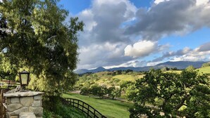 Un sendero de piedra conduce a una valla de madera con vistas a un paisaje verde exuberante, árboles y montañas distantes bajo un cielo parcialmente nublado.