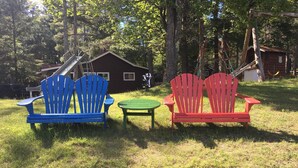Two Adirondack chairs, one blue and one red, placed on a grassy lawn with a wooden cabin in the background. - Beautiful Vacation Home located on Upper Buckatabon Lake (Conover)