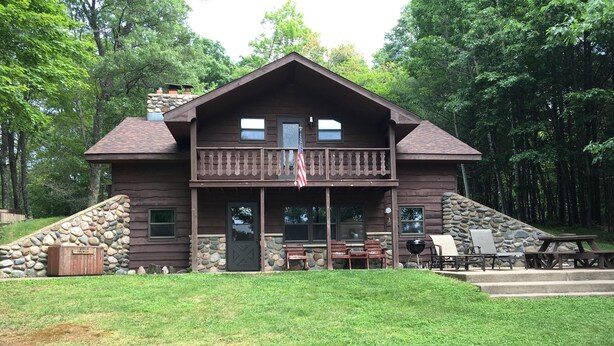 A two-story cabin with a stone foundation, a front porch with wooden railings, and a flag. - Beautiful Vacation Home located on Upper Buckatabon Lake (Conover)