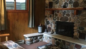 A cozy room with a stone fireplace, wooden furniture, and a window with curtains. - Beautiful Vacation Home located on Upper Buckatabon Lake (Conover)