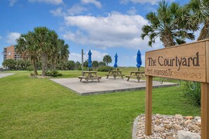 A sign for 'The Courtyard Port Aransas, TX' with picnic tables and umbrellas in a grassy area.