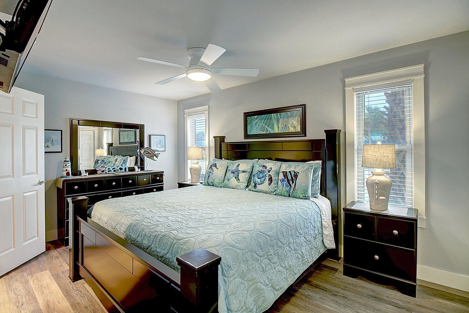 Une chambre à coucher comprenant un lit, une commode, une table de chevet, un ventilateur de plafond et une fenêtre avec des stores.