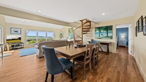 A spacious living area with a wooden dining table, blue upholstered chairs, a wooden staircase, and large windows offering a view of the outdoors. - Waterfront Beach Club Villa! 3 BR/3.5 BA! Luxury, Ocean view! Amenity access included! (Seabrook Island)