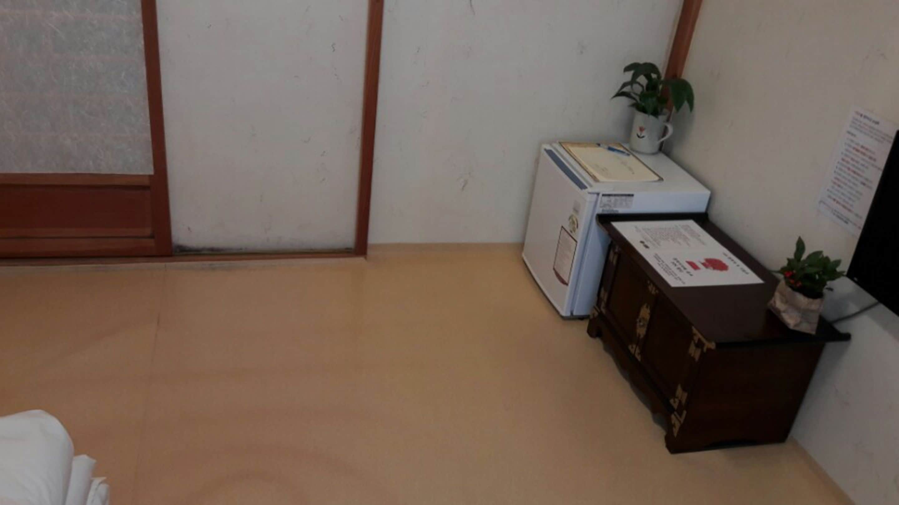 Um quarto com carpete bege, parede branca, porta de madeira, uma pequena mesa branca com uma planta, uma impressora branca e uma cômoda marrom.