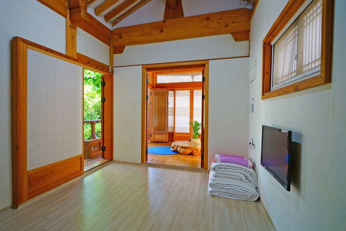 Una habitación con vigas de madera, un esterilla de tatami, un televisor y vistas a un jardín a través de una puerta corredera.