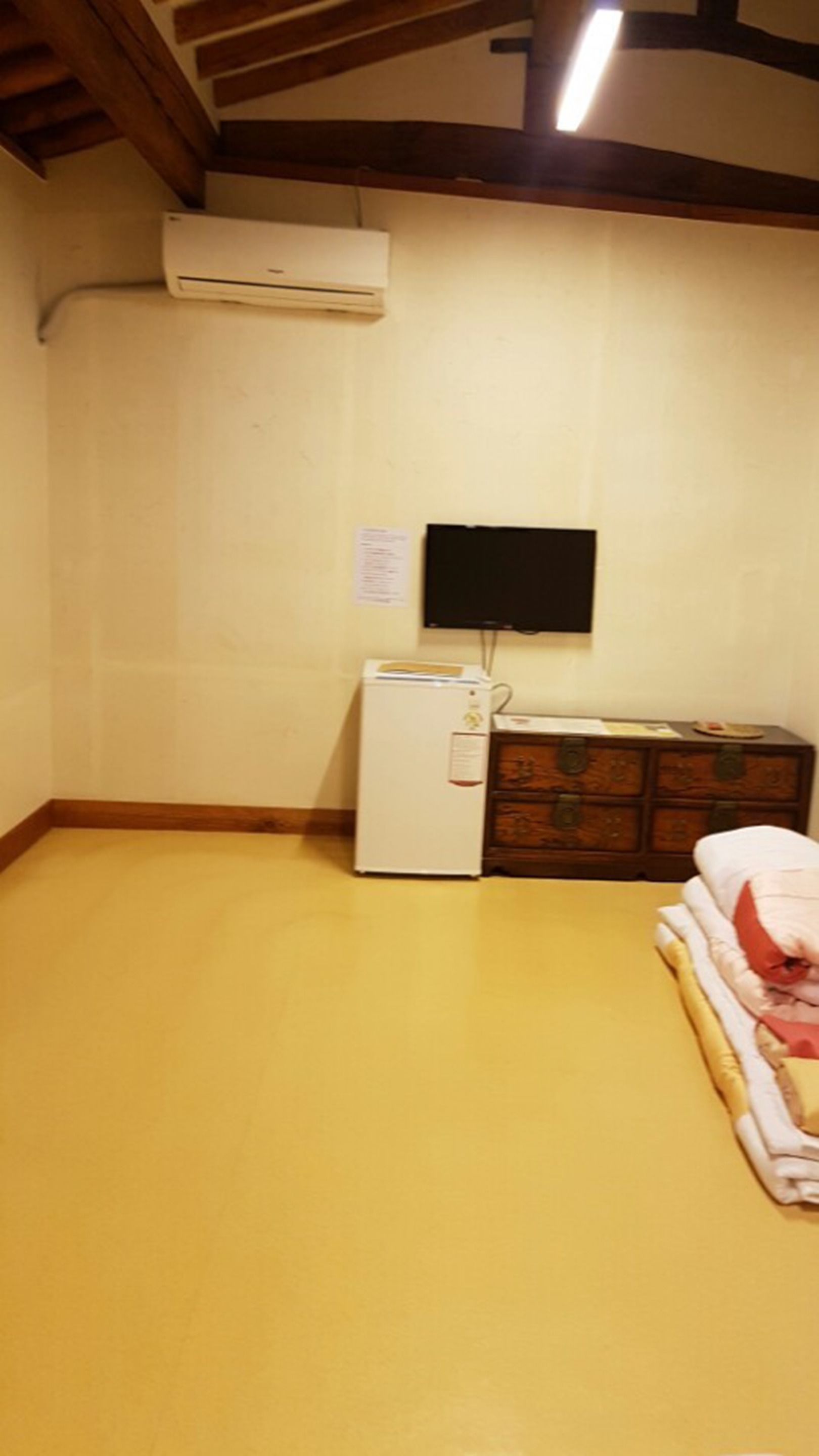 Une pièce avec un plancher jaune, un réfrigérateur blanc, une commode en bois et un téléviseur fixé au mur.