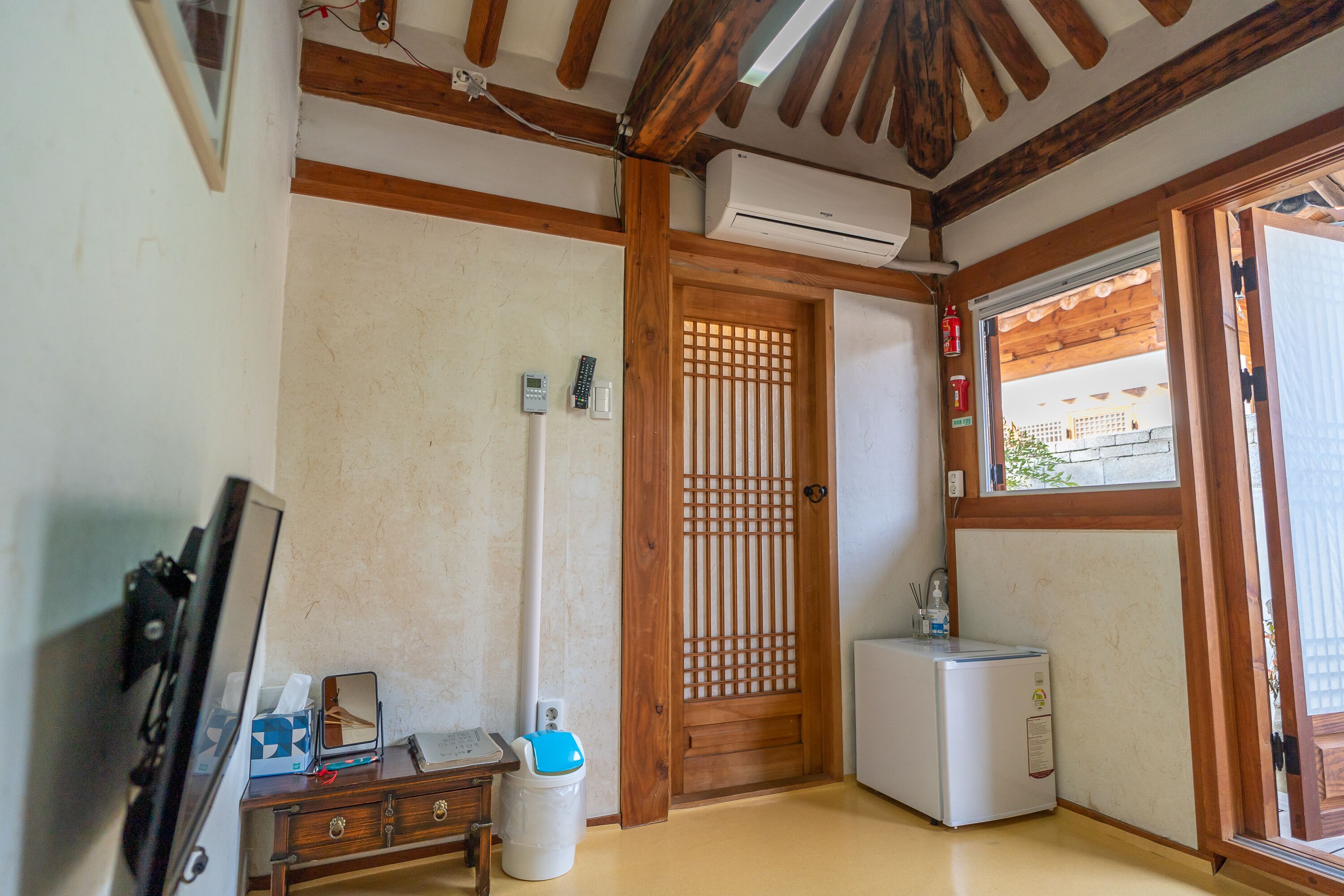 A compact room with a wooden ceiling, a small refrigerator, a heater, a wooden door with a lattice design, and a window with a view of the outside.