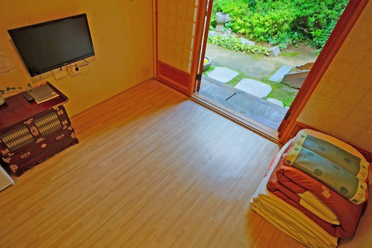 Una habitación pequeña con suelo de madera, un televisor en la pared, un mueble pequeño de madera y una ventana que da a un jardín.