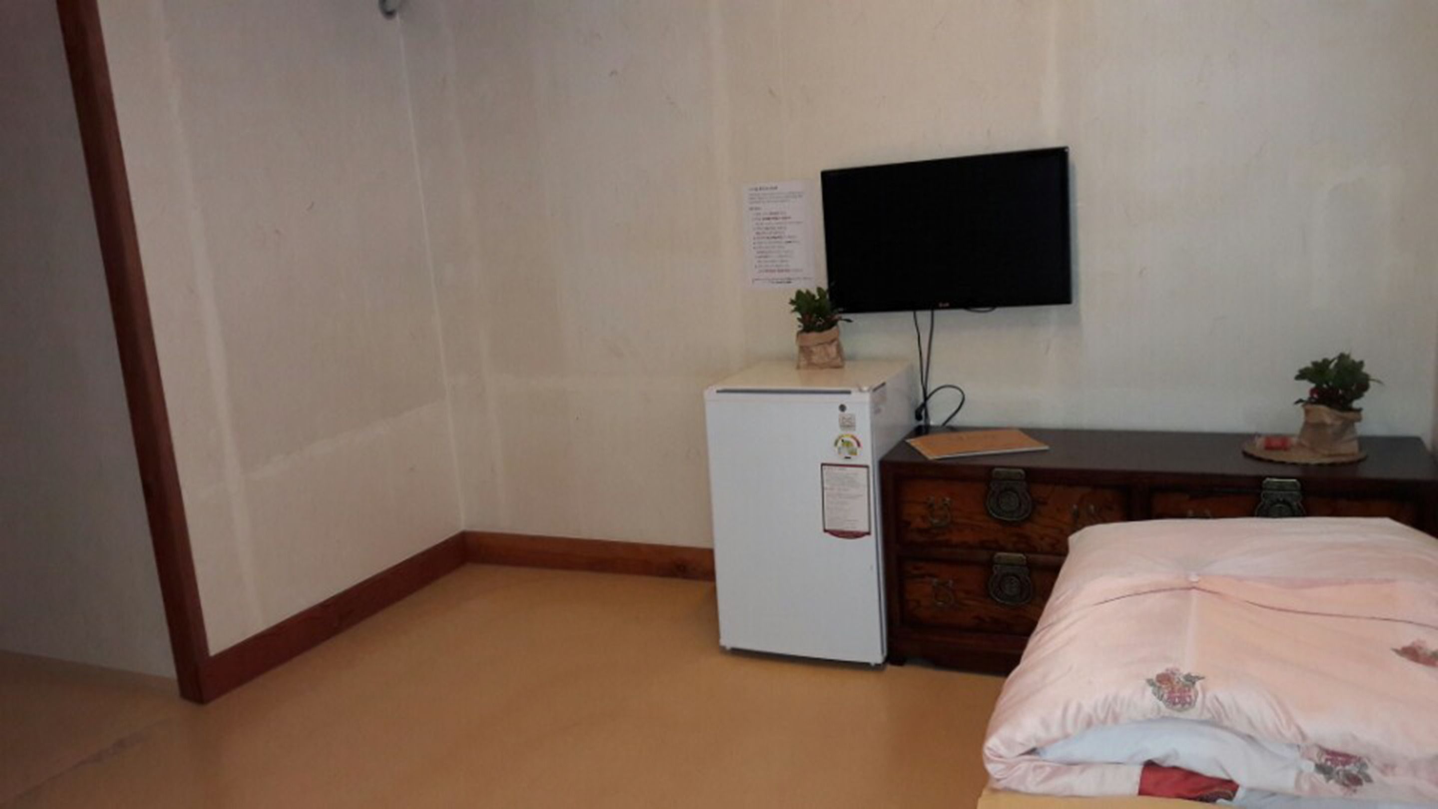 Una habitación pequeña con un refrigerador blanco, un aparador de madera, una cama con una manta rosa y un televisor de pantalla plana montado en la pared.