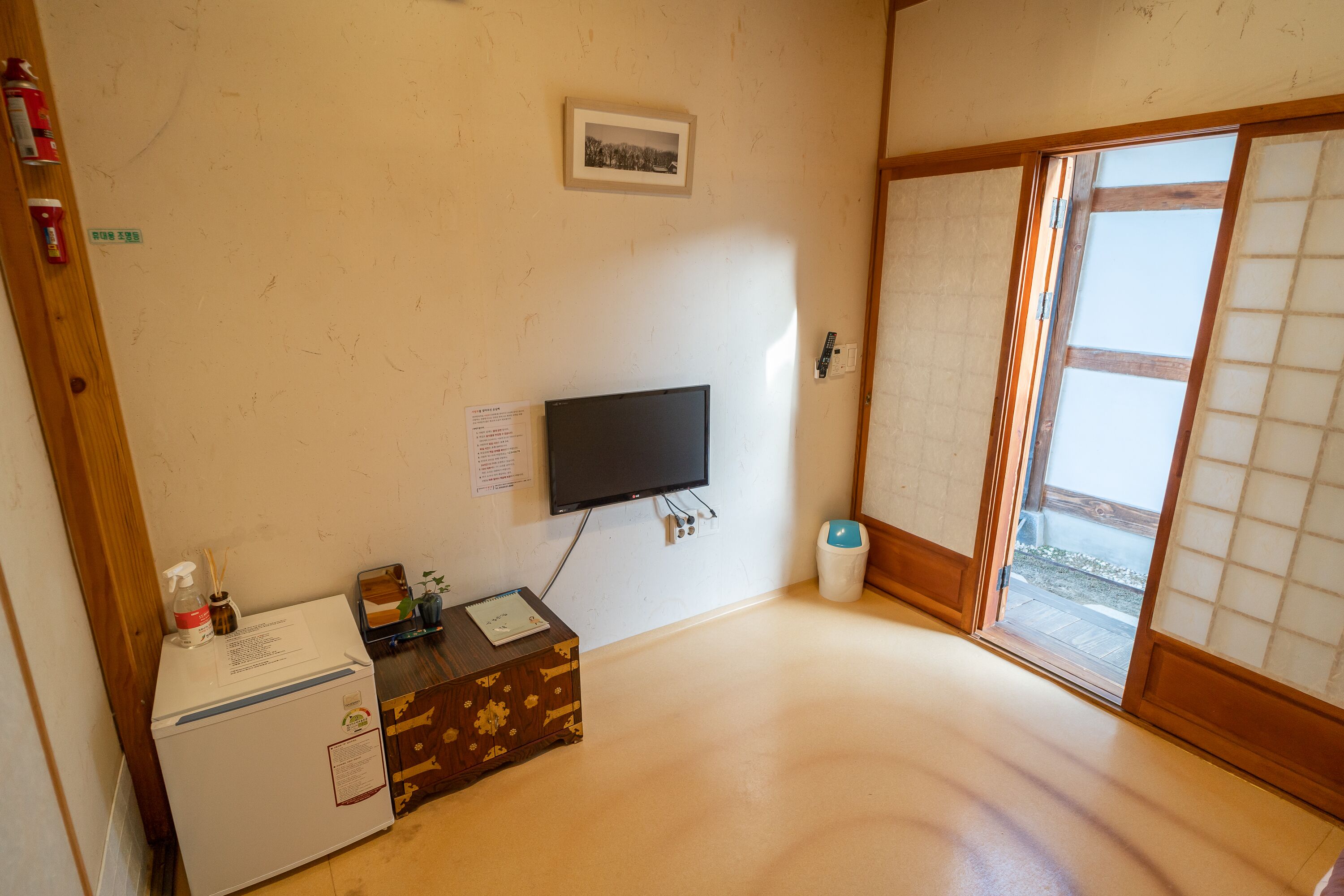 Quarto com televisão, mini-geladeira, armário de madeira, lixeira e porta deslizante com vista externa.
