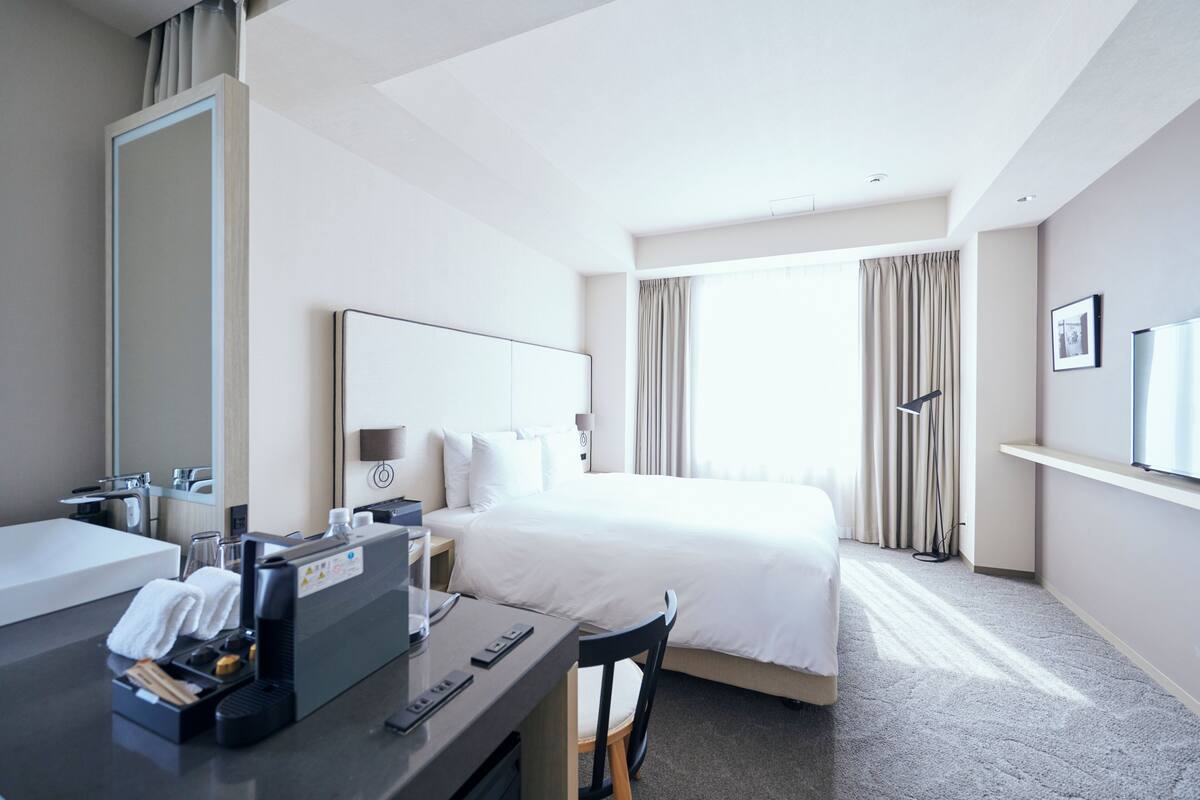 A hotel room with a bed, a desk with a computer, a mirror, and a window with curtains.