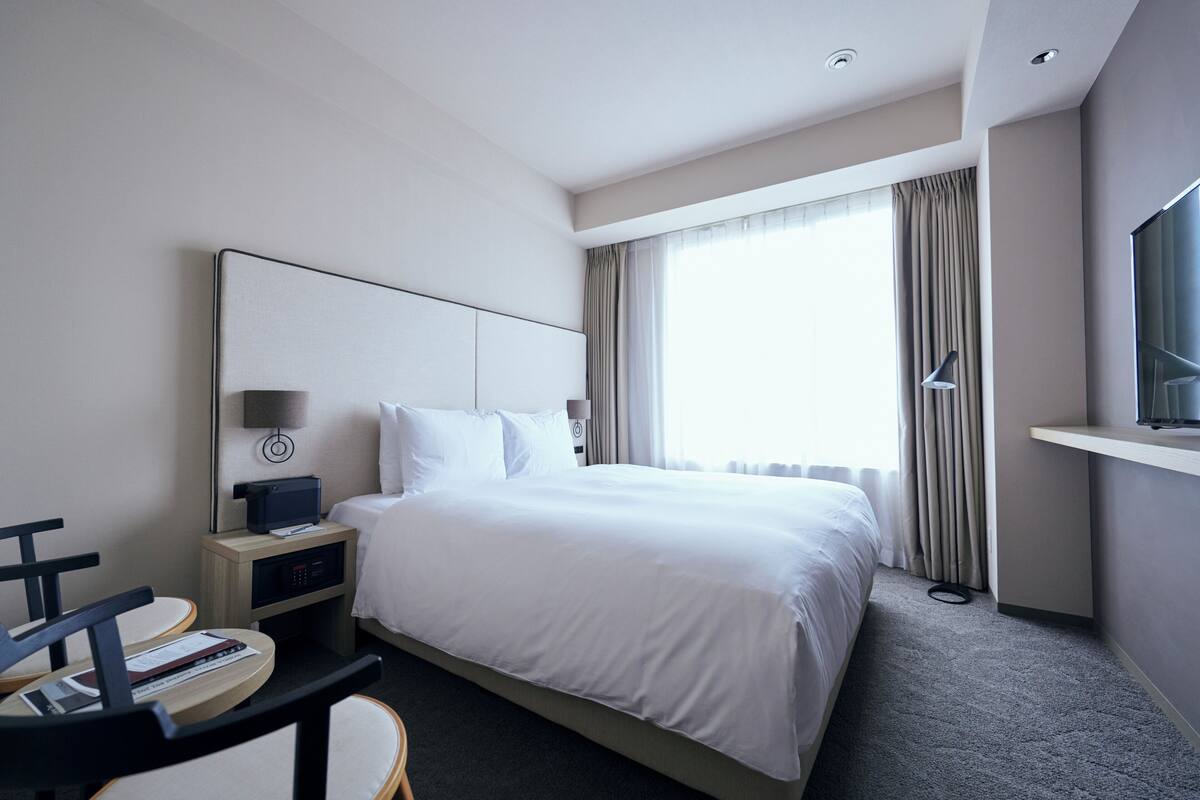 A hotel room with a large bed, a desk with a chair, a flat-screen TV, and a window with curtains.