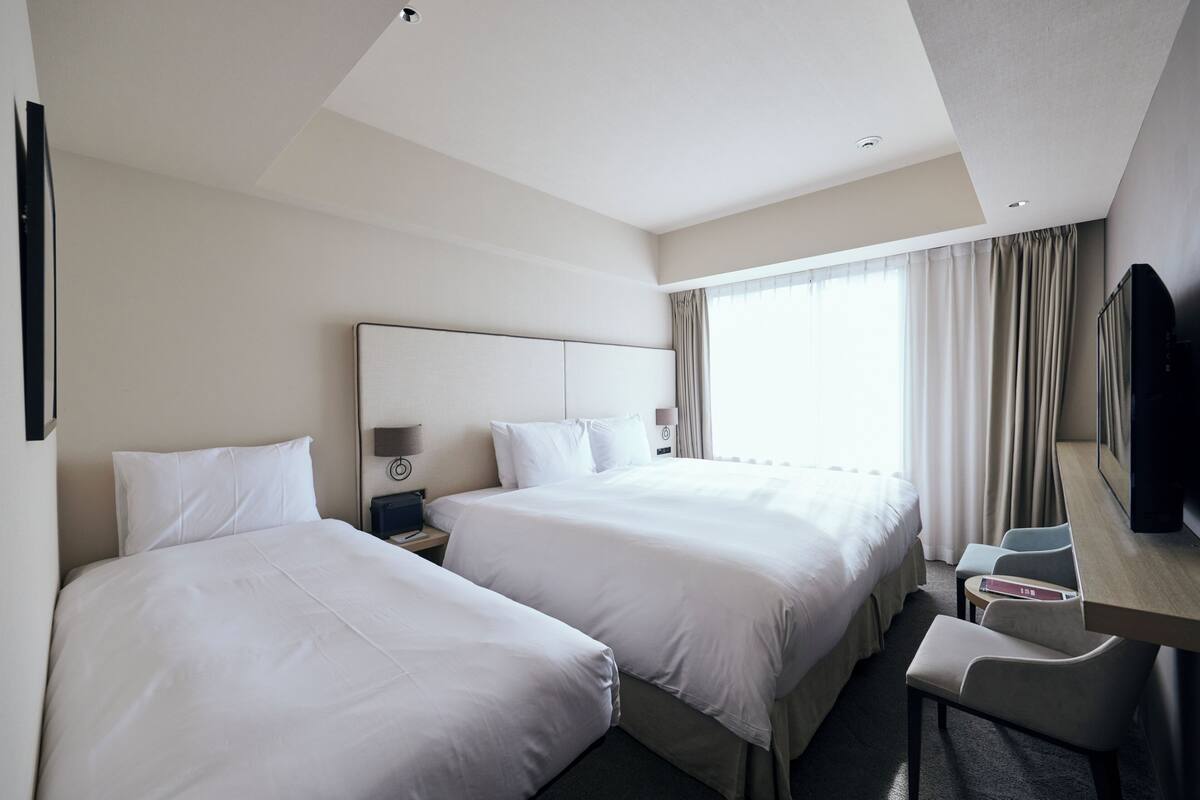 A hotel room with two beds, a desk with a chair, a TV, and a window with curtains.