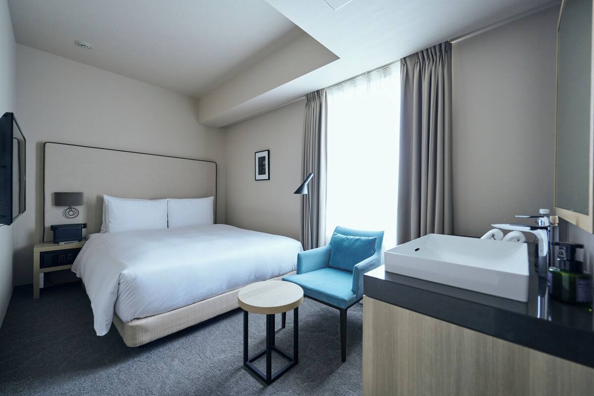A hotel room with a bed, a desk, a chair, a round table, a bathroom with a sink and mirror, and a window with curtains.