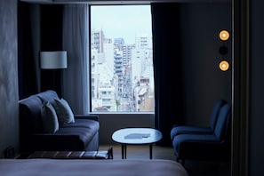 A hotel room with a bed, a sofa, a small table, and a view of a cityscape through a large window.
