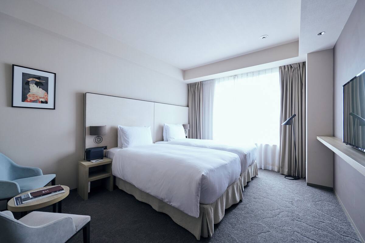 A hotel room with two beds, a desk, a chair, a TV, and a large window with curtains.