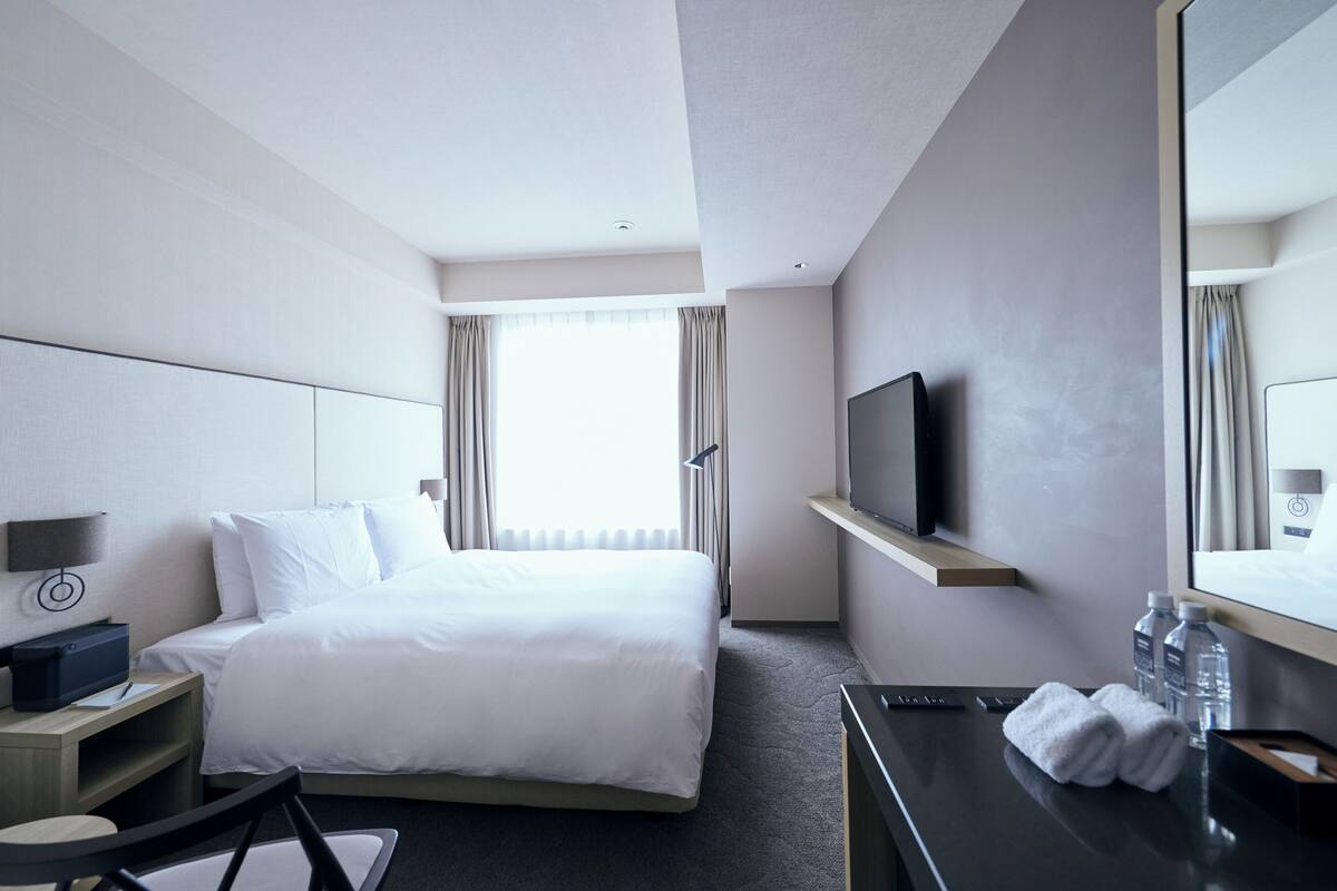 A hotel room with a bed, a TV mounted on the wall, a desk with a chair, and a window with curtains.